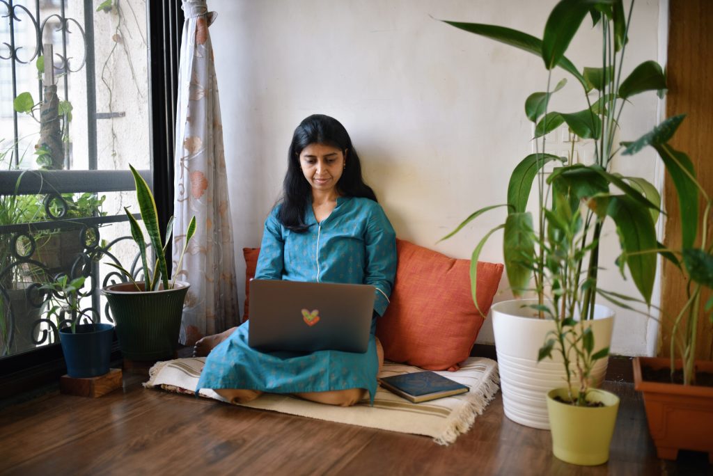 Woman working on laptop sitting next to window surrounded with plant pots